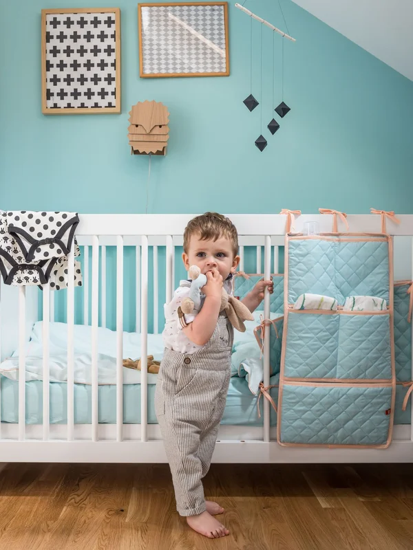  Dziecko stojące przy białym łóżeczku niemowlęcym z pastelowym organizerem na akcesoria.
